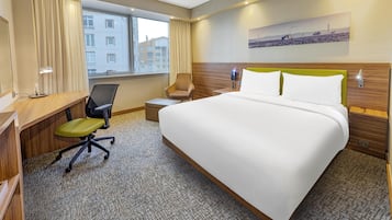 Ett hotellrum med en stor säng, ett skrivbord med en stol, ett litet bord, ett fönster med gardiner och en tavla som hänger på väggen.