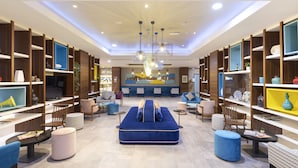 A modern hotel lobby with a central seating area, wooden shelving with decorative items, and a large mural on the back wall.