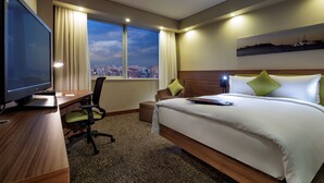 A hotel room with a bed, a desk with a TV, a chair, a window with a city view, and a painting on the wall.