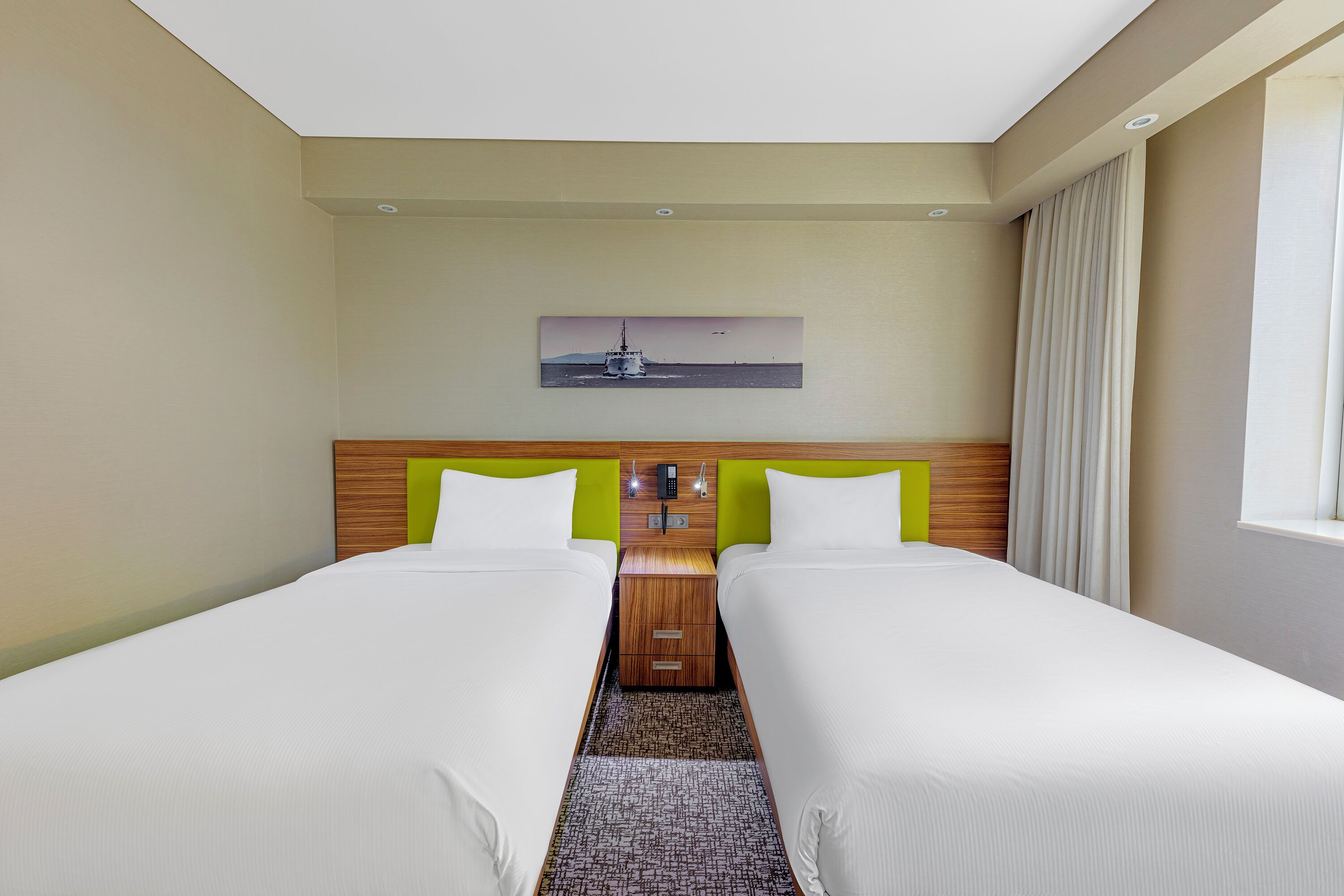 A hotel room with two beds, a wooden headboard, a painting above the beds, and a window with curtains.