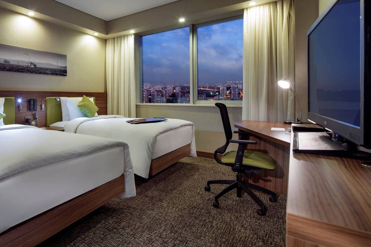 Habitació d'hotel amb dos llits, una escriptori, una cadira, un televisor i un finestral gran amb vista a la ciutat.