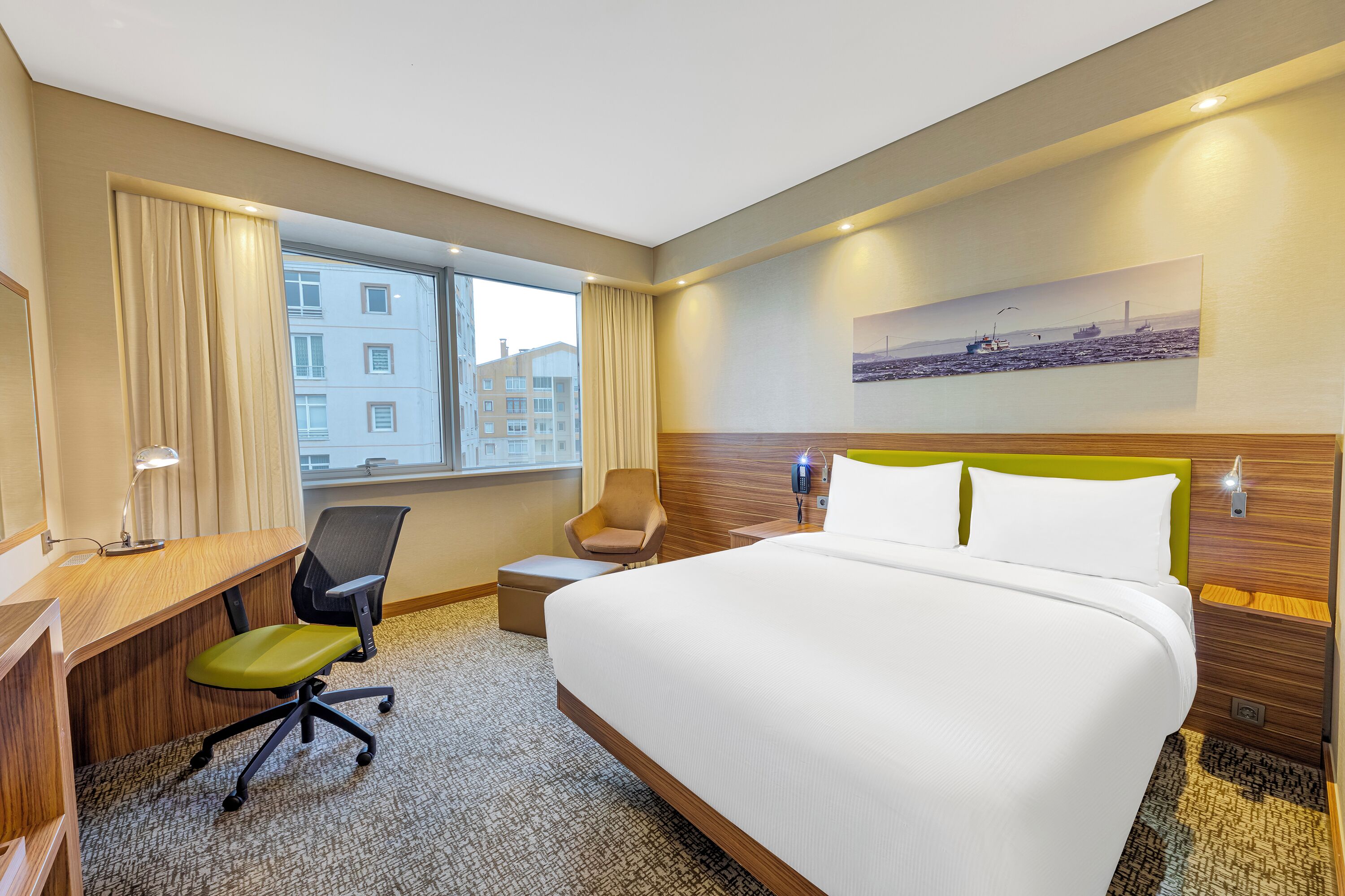 A hotel room with a bed, desk, chair, and a large window.