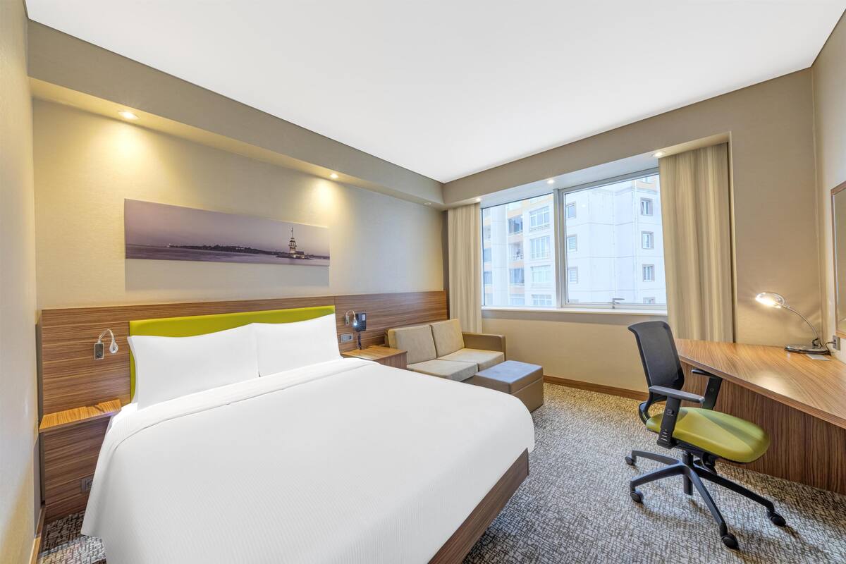 Habitació d'hotel amb un llit gran, una taula de treball, una cadira, un sofà i una finestra amb vista a edificis.