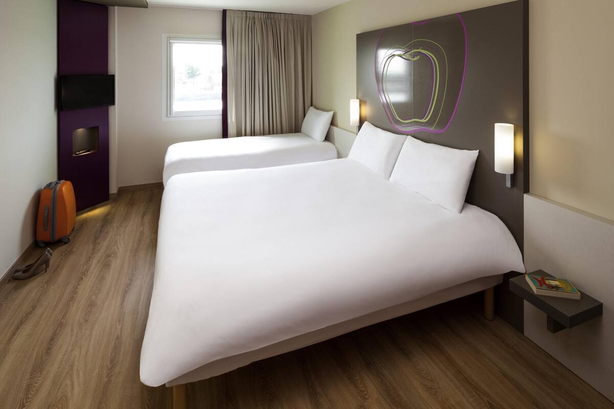 A hotel room with two beds, a TV, a window with curtains, a wall art piece, and a bedside table with a book.