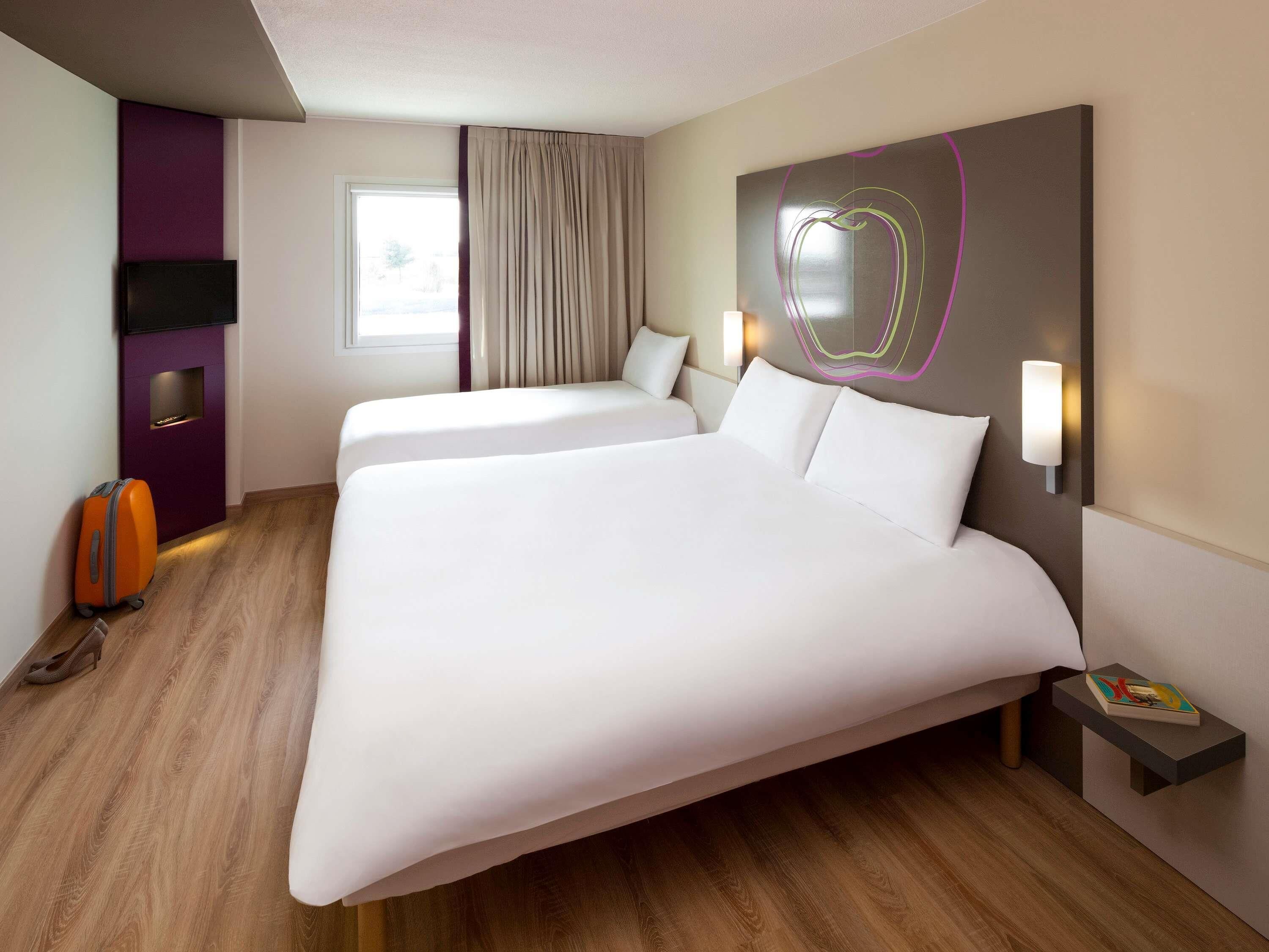 Ein Hotelzimmer mit zwei Betten, einem Flachbildfernseher, einem Nachttisch mit einem Buch und einem Fenster mit Vorhängen.