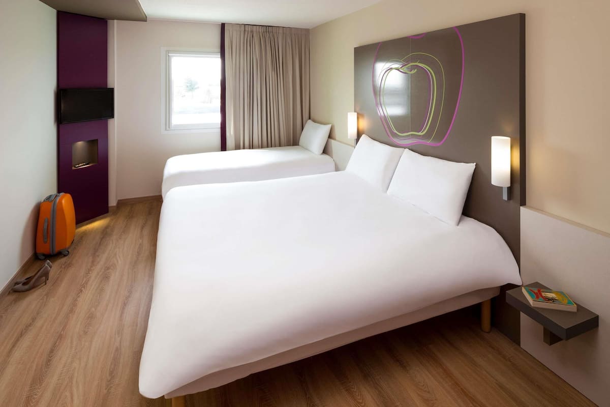 Ein Hotelzimmer mit zwei Betten, einem Flachbildfernseher, einem Nachttisch mit einem Buch und einem Fenster mit Vorhängen.