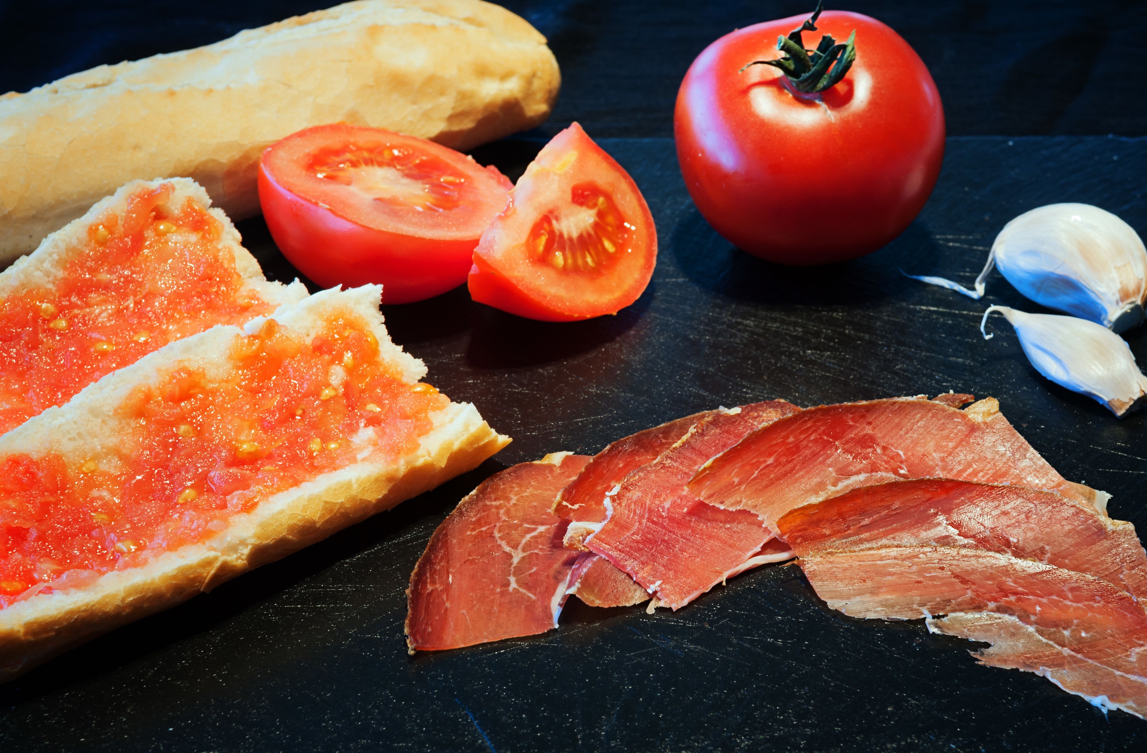 Una baguette, rodajas de tomate, un tomate entero y ajo en rodajas sobre una superficie oscura.