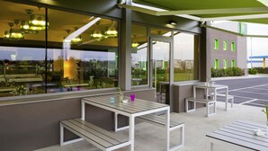 An outdoor seating area with tables and benches, large windows, and a building in the background. - ibis Styles Lleida Torrefarrera (Torrefarrera)