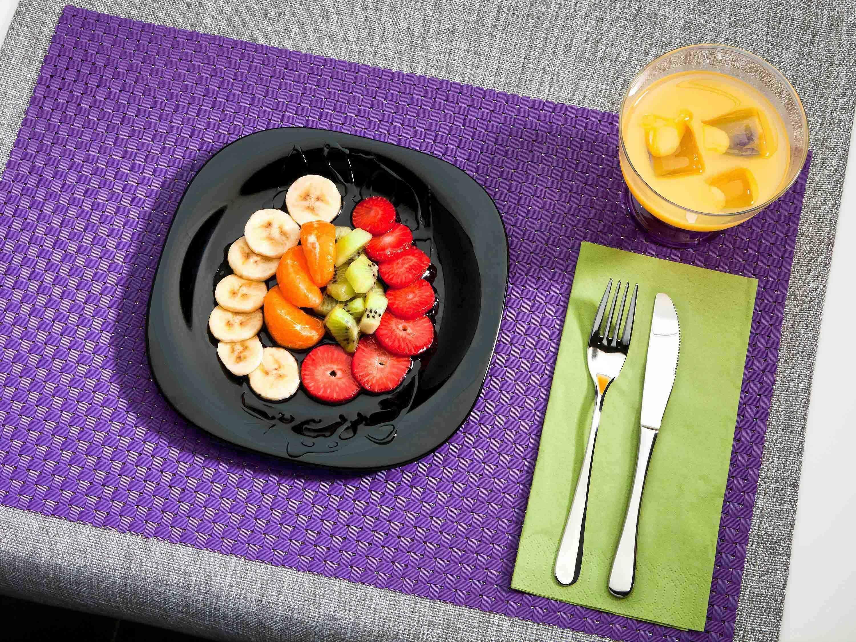 A plate of sliced fruits on a purple mat, a green napkin with cutlery, and a glass of orange juice.