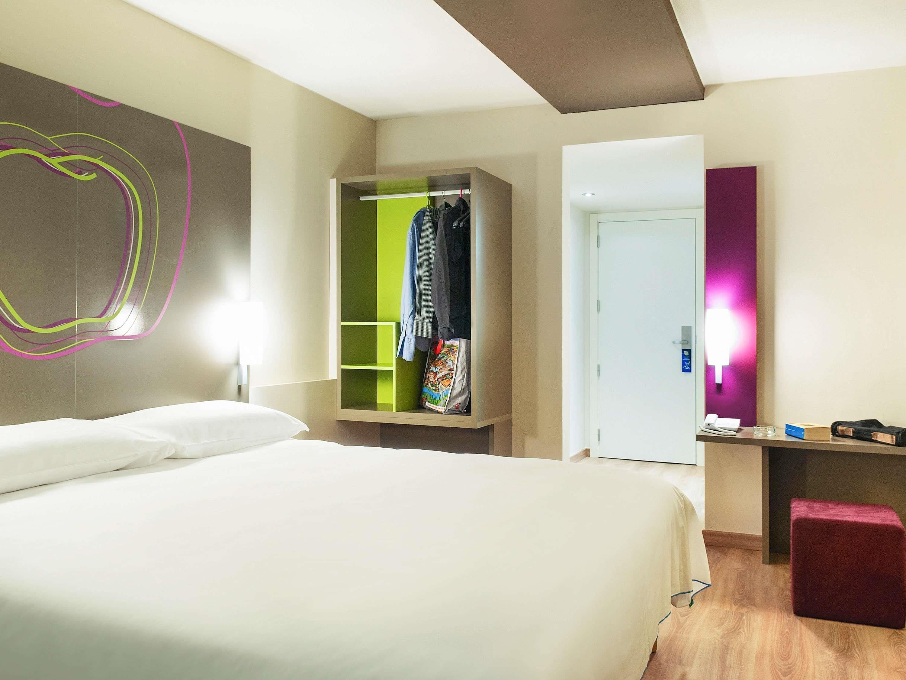 Hotellihuone, jossa on sänky, vaatekaappi, työpöytä tuolilla ja ovi.