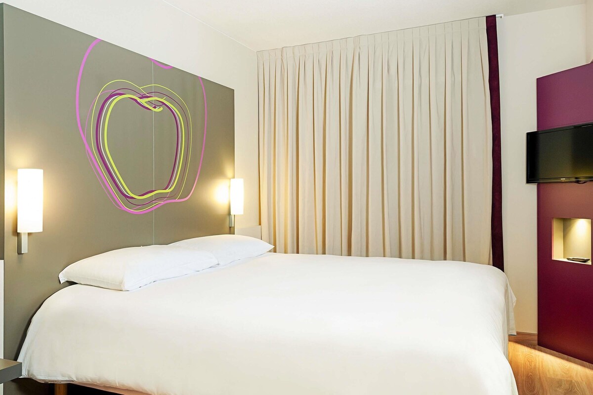 Habitación de hotel con cama, televisor montado en la pared y una lámpara decorativa.