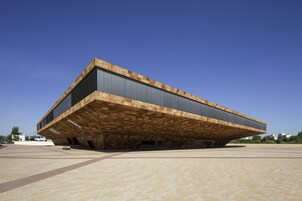 Un edificio moderno con un diseño cantilever único, ventanas amplias y una plaza espaciosa al frente.