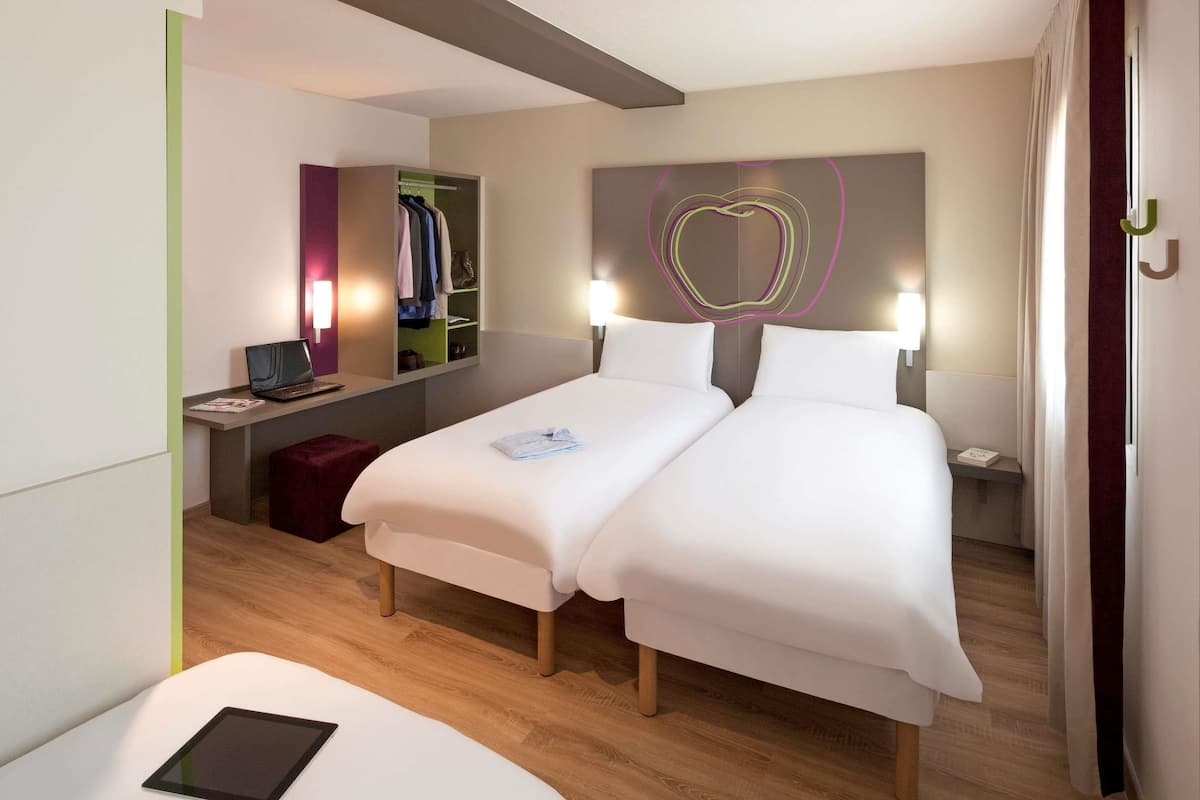 A modern hotel room with a double bed, bedside tables, a desk with a laptop, a red ottoman, and a tablet on a white couch.