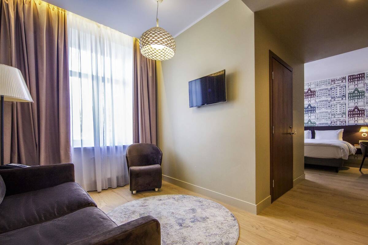 Une chambre d’hôtel comprenant un lit, une télévision, une chaise, un tapis rond et une porte donnant sur une autre pièce.