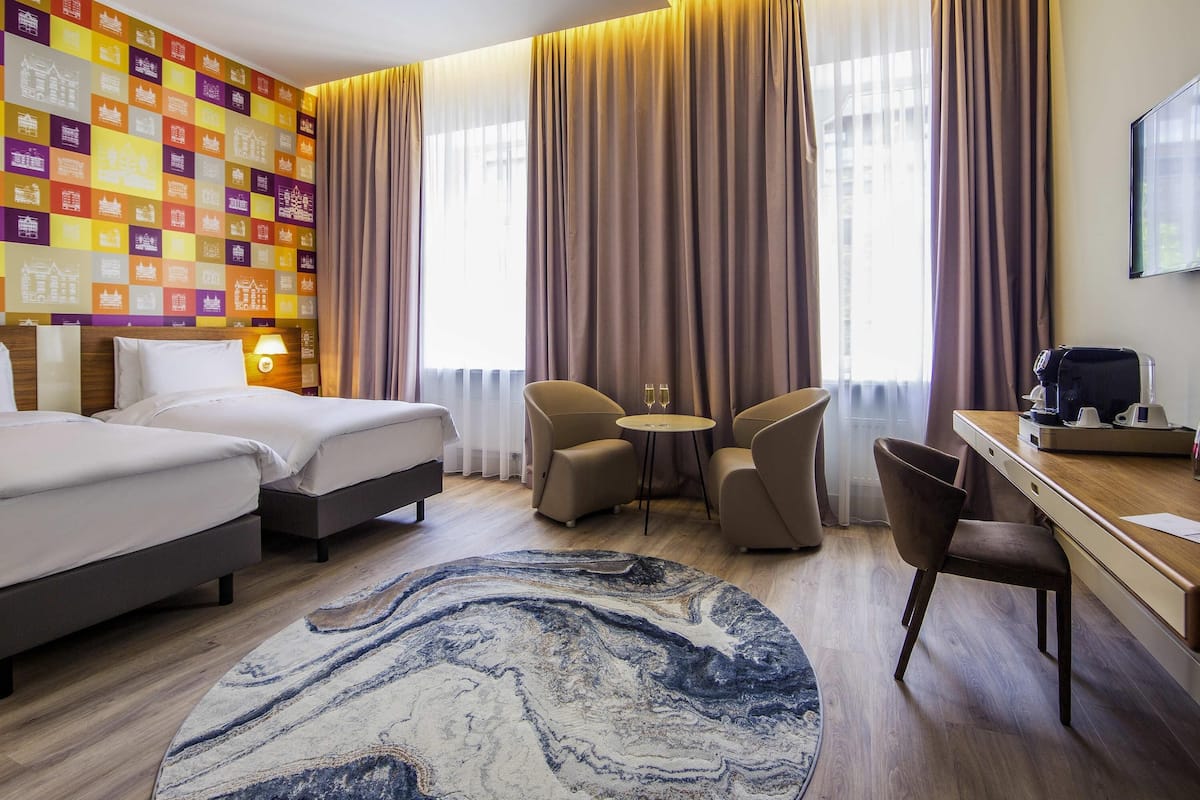 Une chambre d’hôtel avec deux lits, un bureau avec une machine à café, une petite table avec deux verres de vin et une grande fenêtre avec des rideaux.