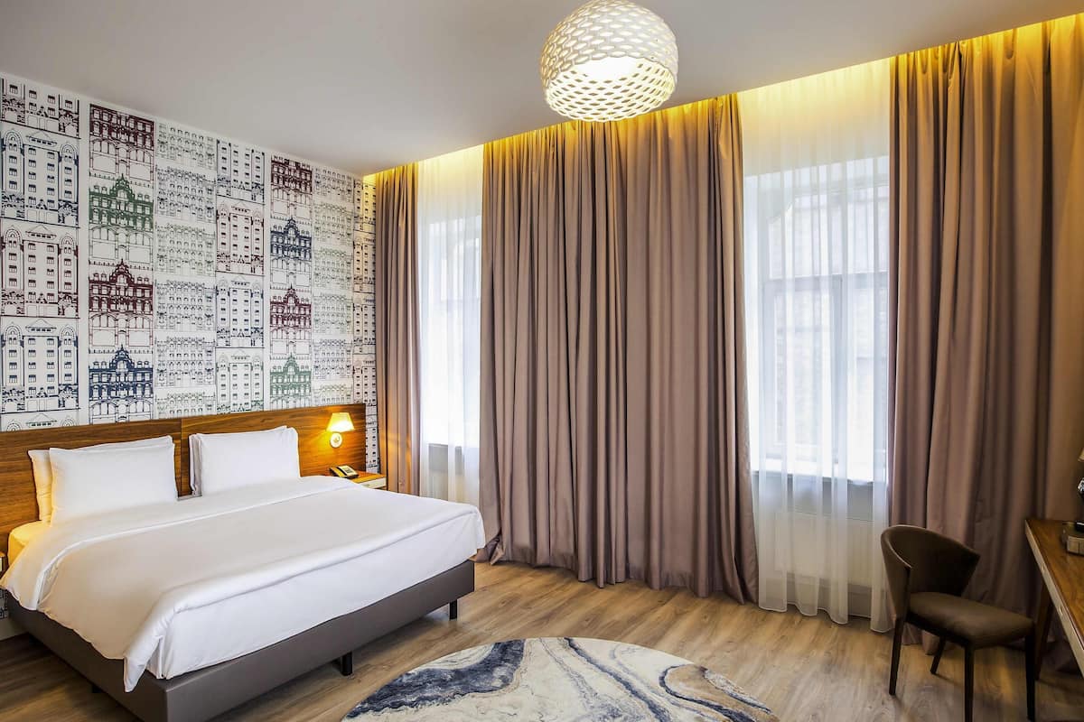 Une chambre d’hôtel avec un grand lit, un tapis rond, un bureau avec une chaise et un mur recouvert d’un papier peint représentant un paysage urbain.