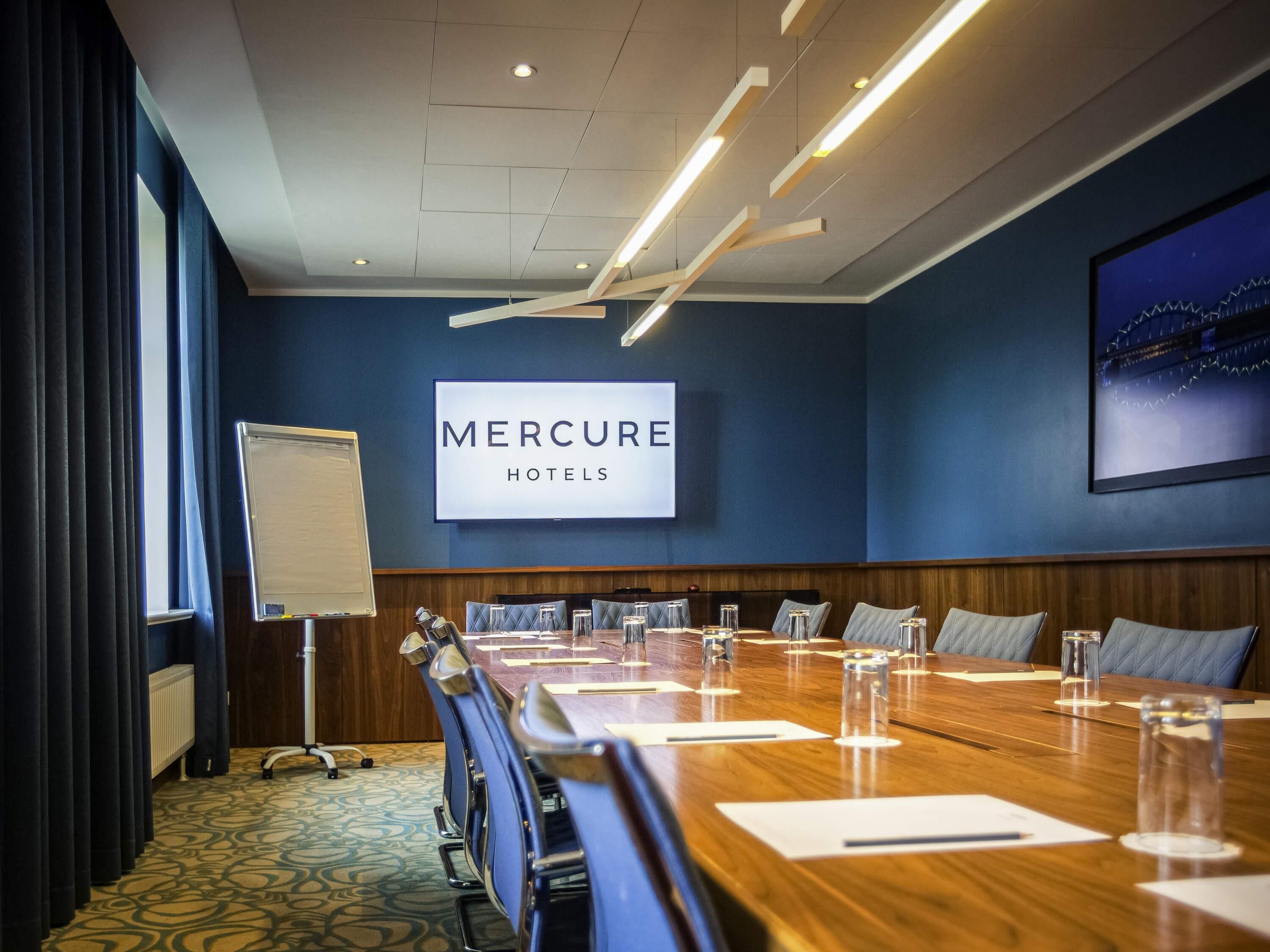 A conference room with a long table, chairs, a whiteboard, and a sign reading 'Mercure Hotels'.