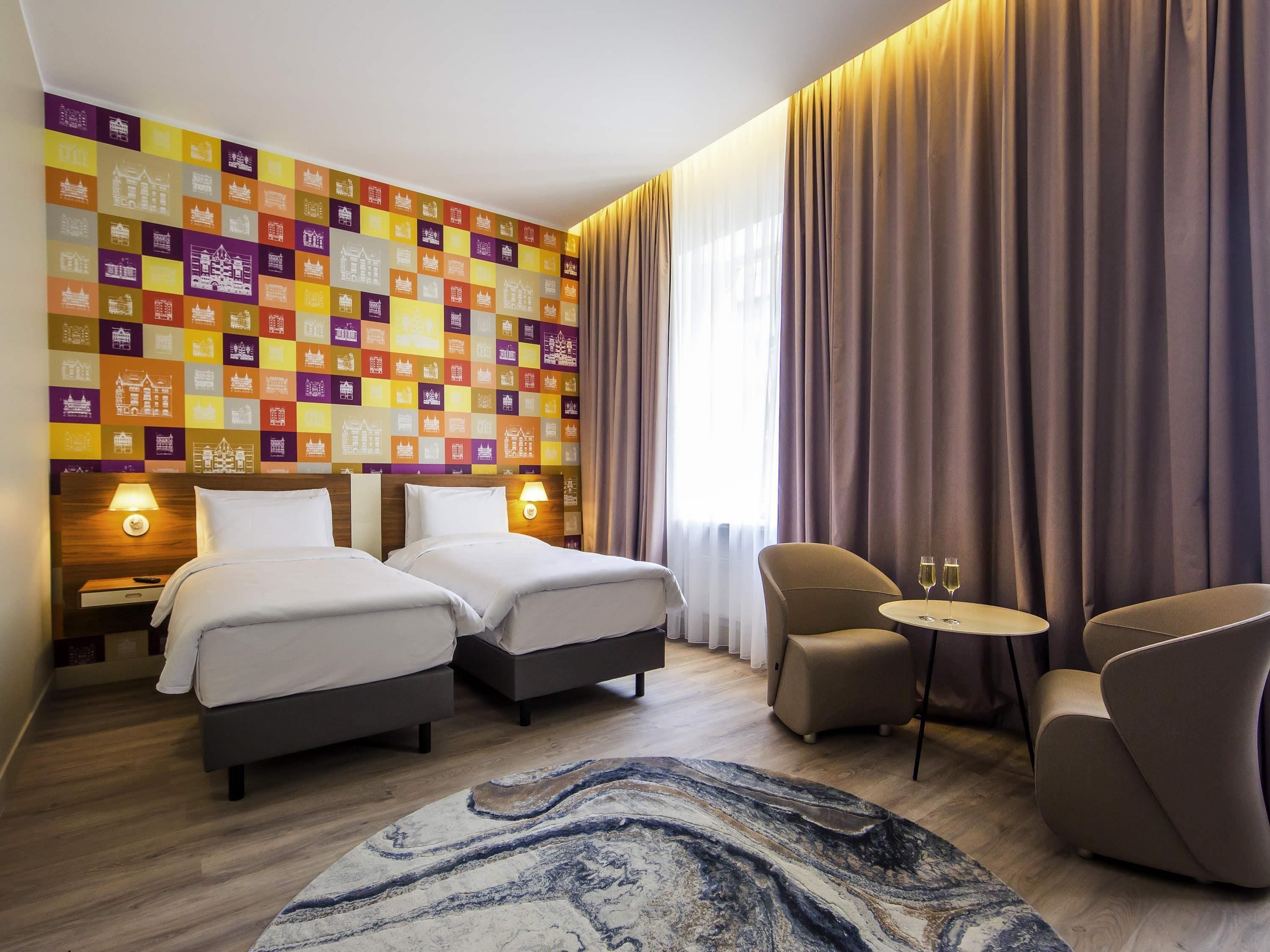 Habitación de hotel con dos camas, un área de estar con una mesa y una pared decorada con iconos de edificios coloridos.