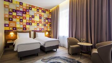 Habitación de hotel con dos camas, un área de estar con una mesa y una pared decorada con iconos de edificios coloridos.