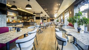A modern restaurant interior with a long counter, multiple tables, and chairs, and a kitchen area in the background. - Mercure Riga Centre (Riga)