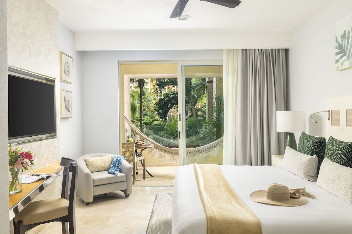 A hotel room with a bed, a desk, a chair, a TV, and a view of a garden through a glass door.