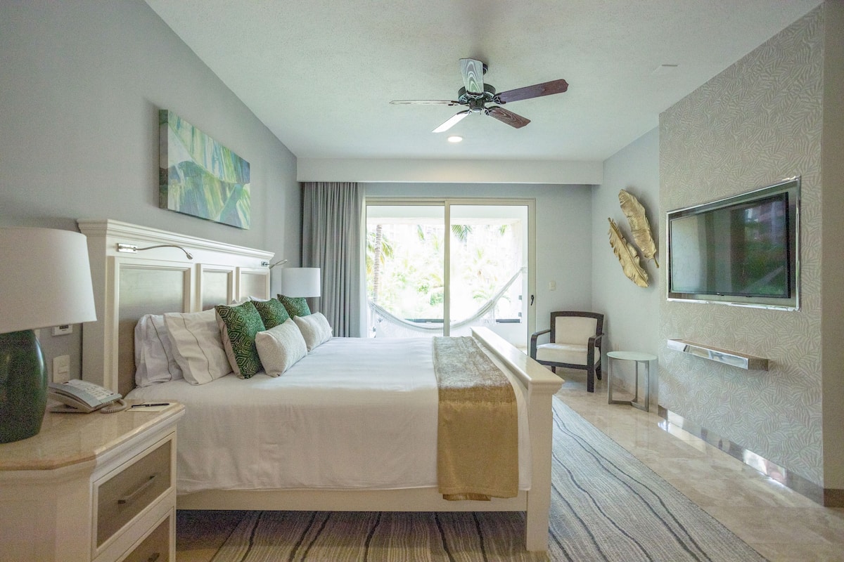 A hotel room with a bed, bedside tables, a ceiling fan, a TV mounted on the wall, and a large window with a view of greenery.