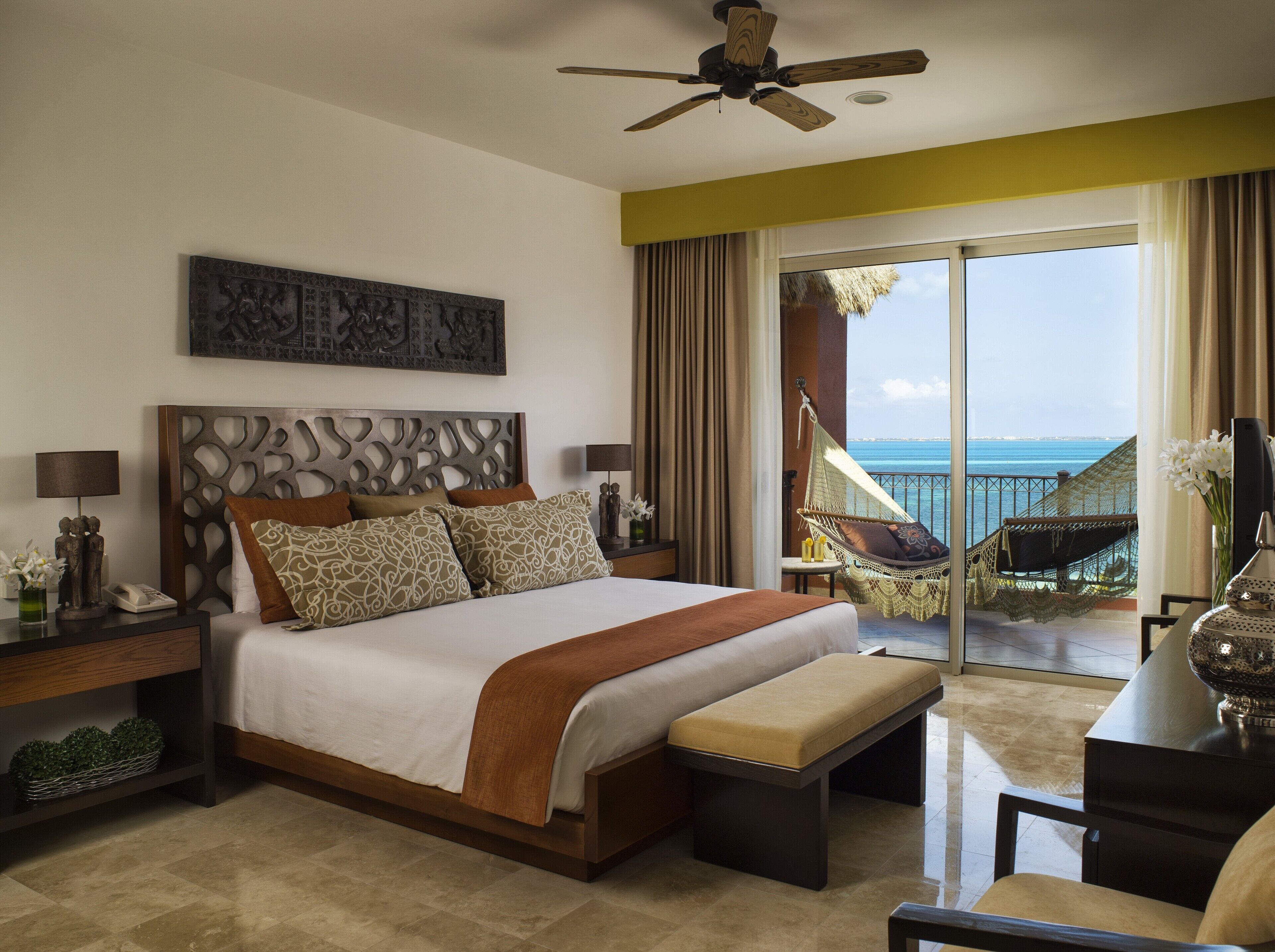 A hotel room with a bed, a nightstand, a desk, a chair, a balcony with a view of the sea, and a ceiling fan.