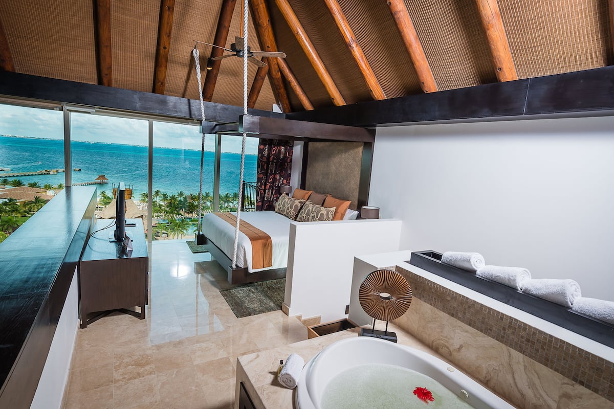 A modern bedroom with a large window overlooking the ocean, a private balcony, and a spacious bathroom with a bathtub and towel rack.