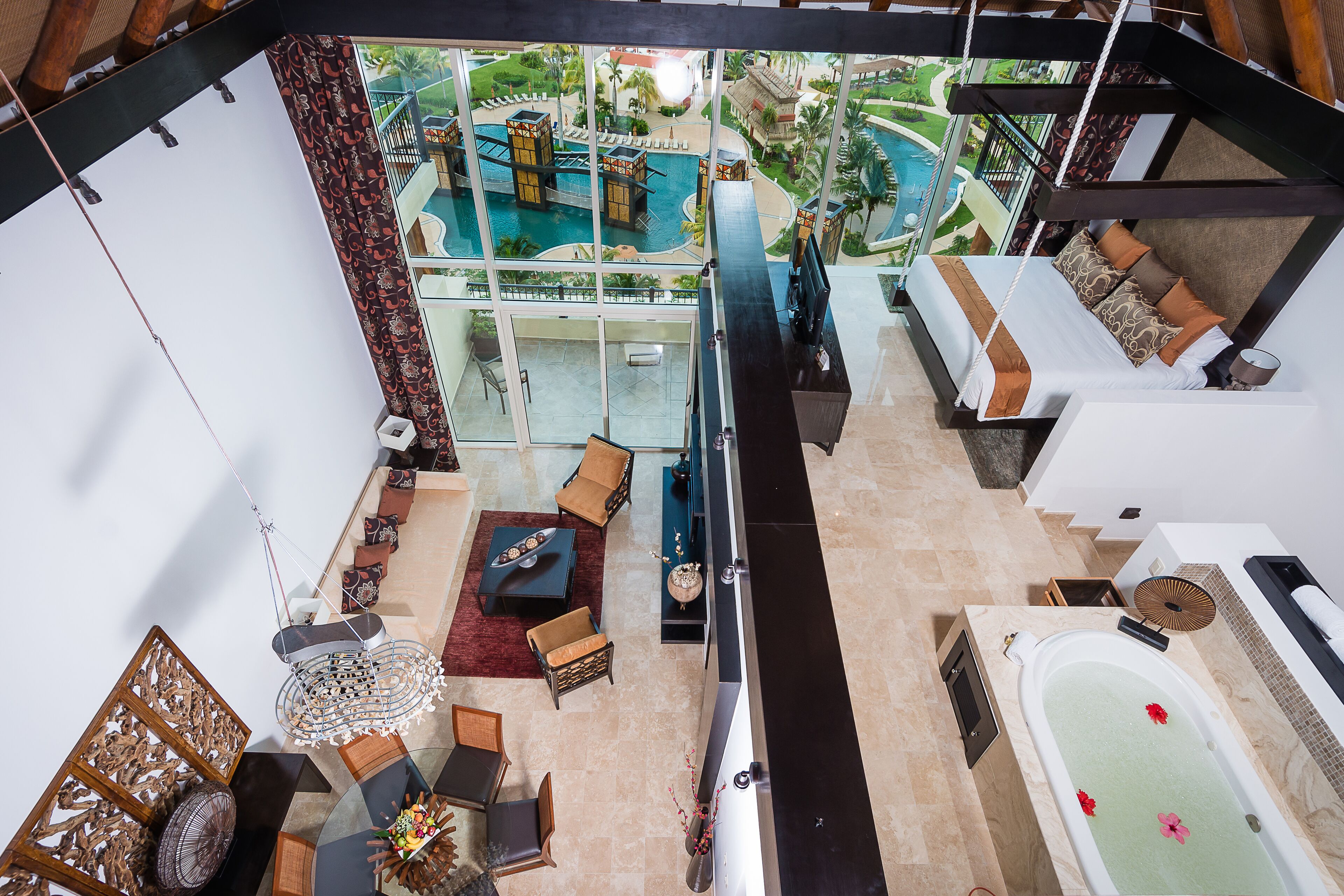 A high-ceilinged room with a large window overlooking a pool area, a sitting area with a coffee table, and a separate section with a bathtub and towels.