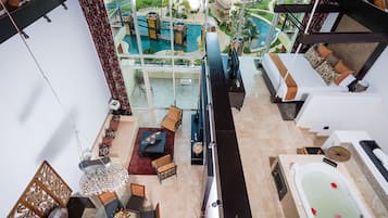 Une pièce aux hauts plafonds, dotée d’une grande fenêtre donnant sur un espace piscine, d’un coin salon avec une table basse et d’un espace séparé comprenant une baignoire et des serviettes.