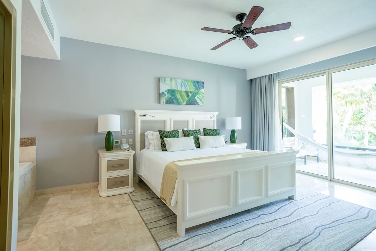 A bedroom with a large bed, white nightstands, a ceiling fan, and a view of the outdoors.