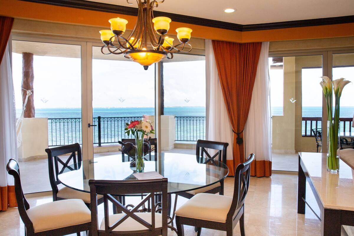 A dining area with a glass table, four chairs, a chandelier, and a view of the sea.