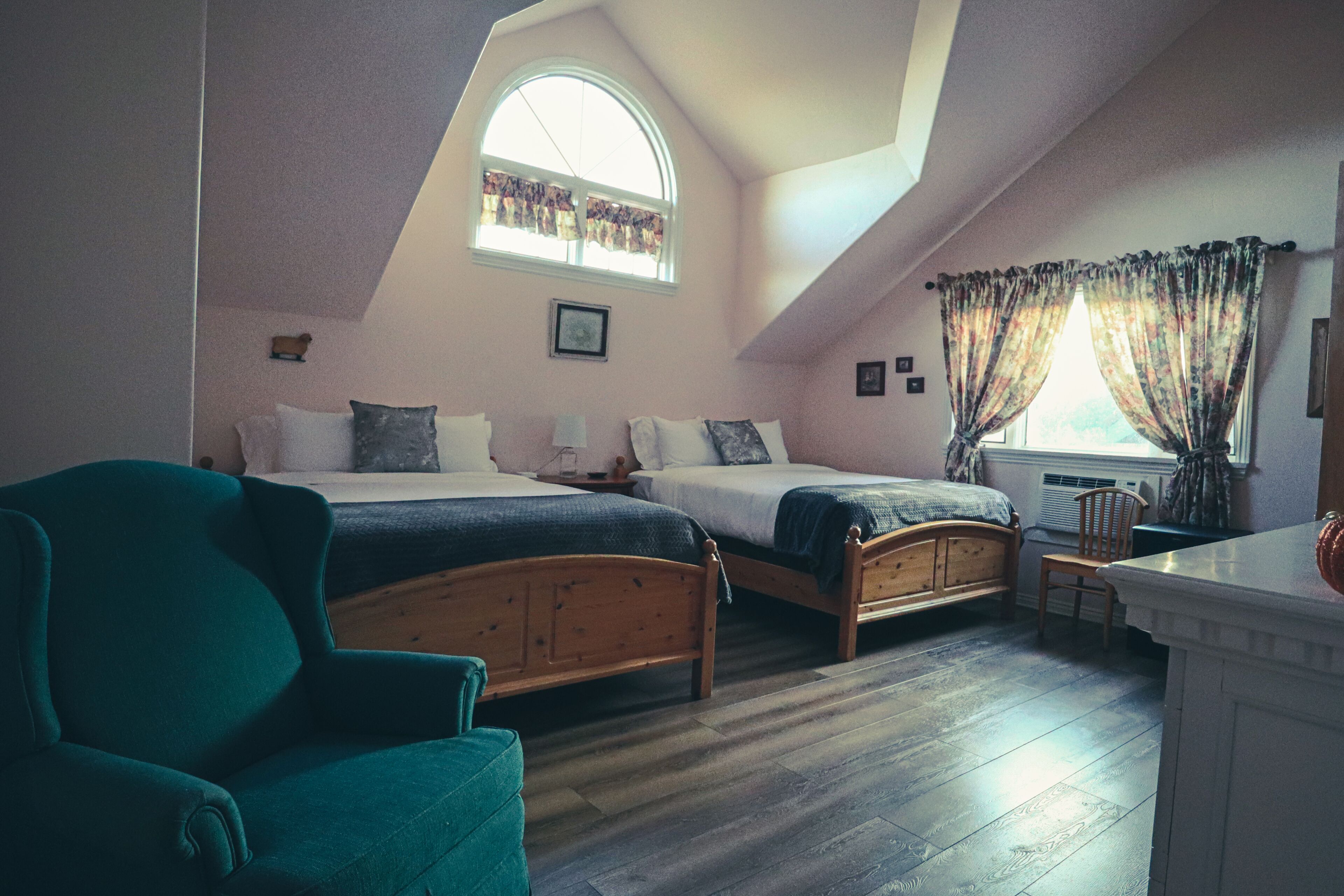 A bedroom with two wooden beds, a green armchair, a window with curtains, and a small table.