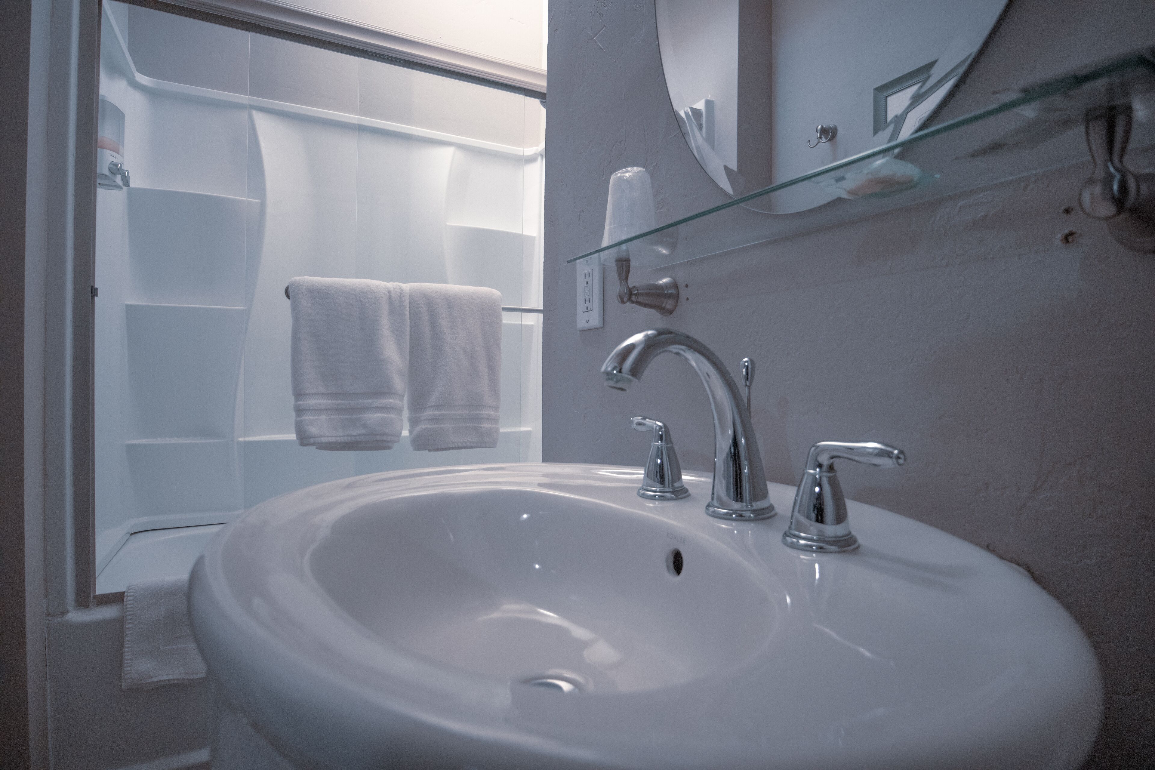 Un baño con un lavamanos blanco, dos grifos y un estante de vidrio sobre el lavamanos. Hay dos toallas blancas colgadas en ganchos.
