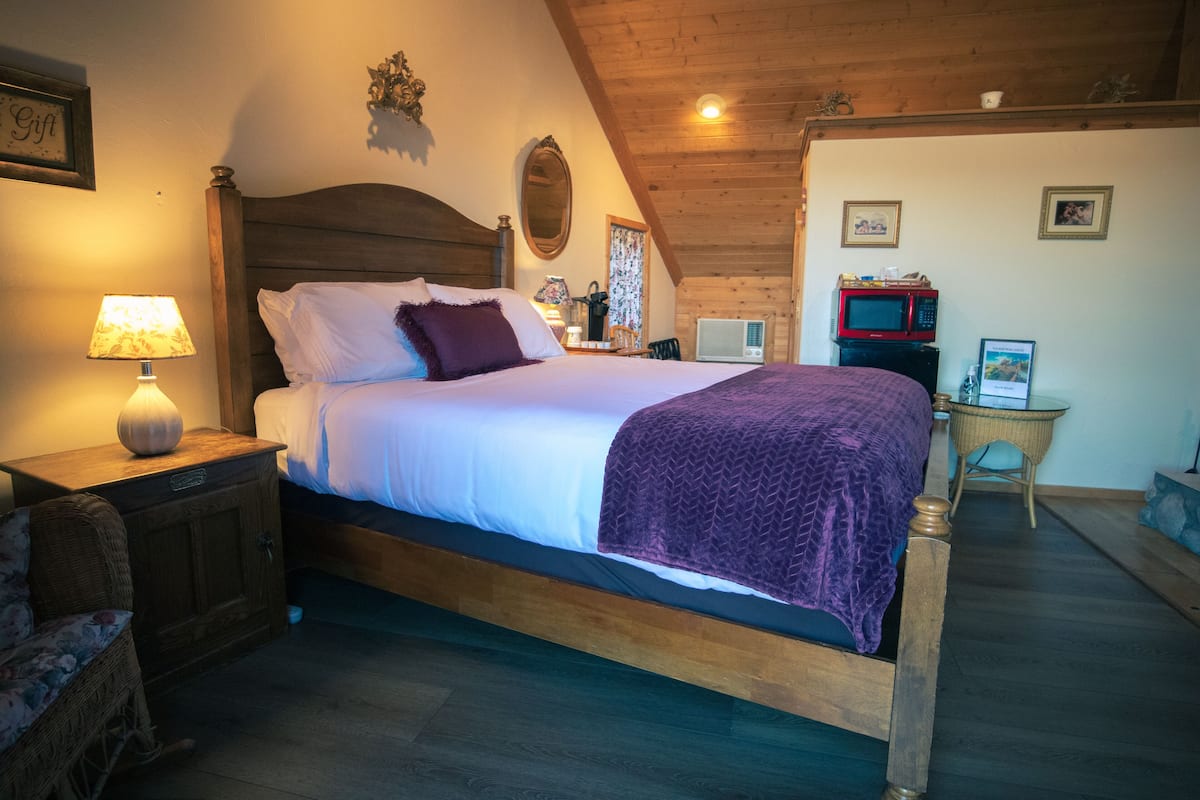 A bedroom with a wooden bed, a nightstand with a lamp, a wicker chair, a small table with a chair, a microwave, and a framed picture on the wall.