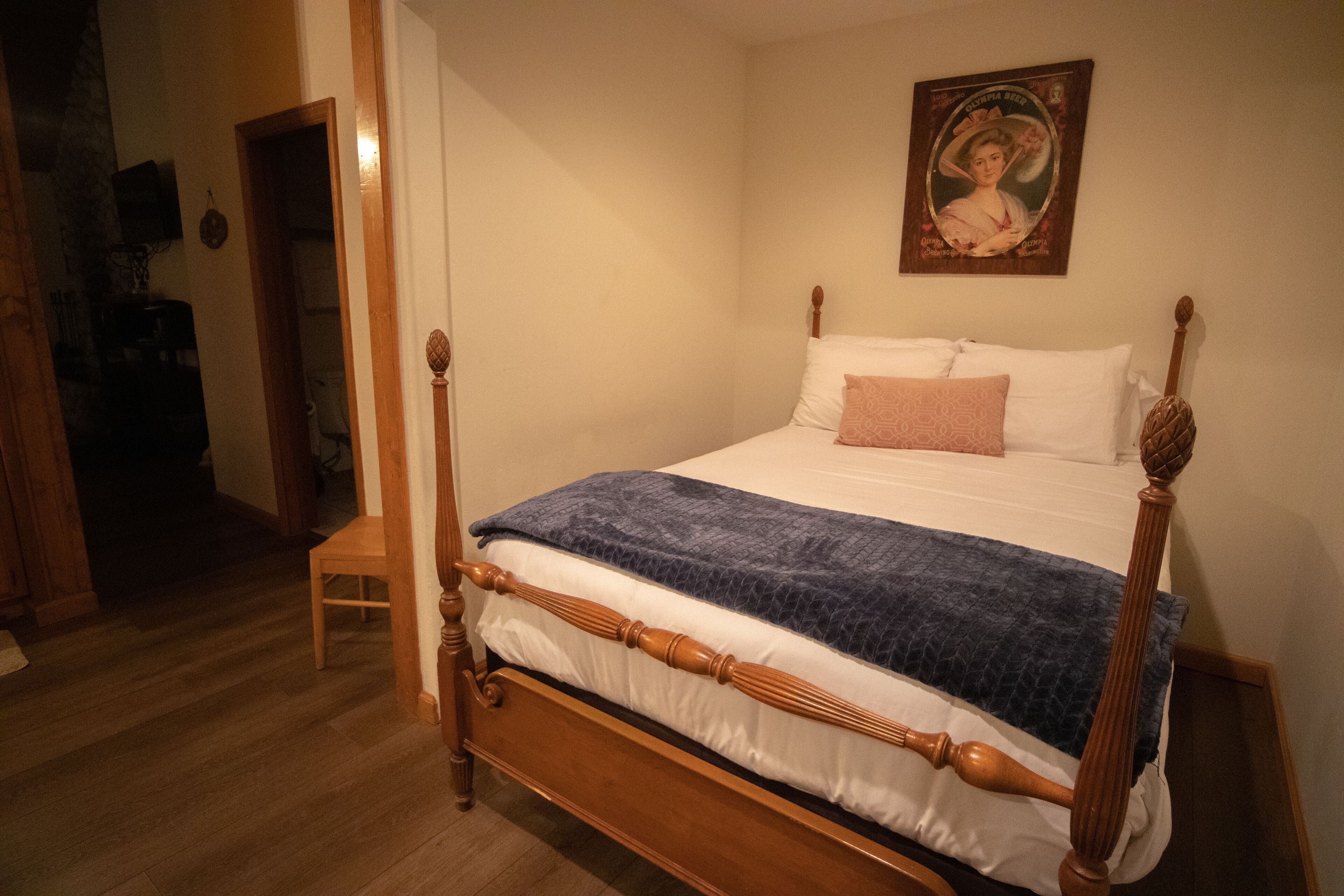A bedroom with a wooden four-poster bed, a single chair, and a portrait on the wall.