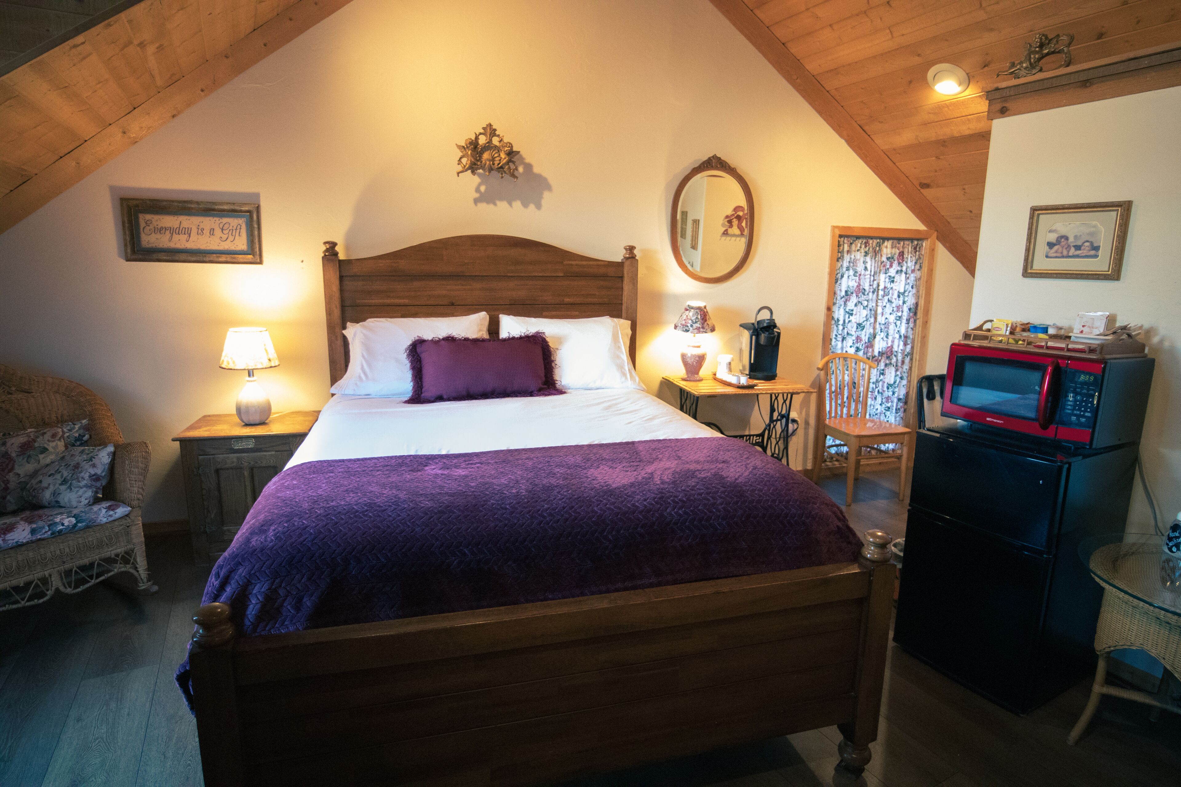 A cozy bedroom with a wooden bed, a nightstand with a lamp, a small table with a lamp, a chair, a microwave, and a refrigerator.