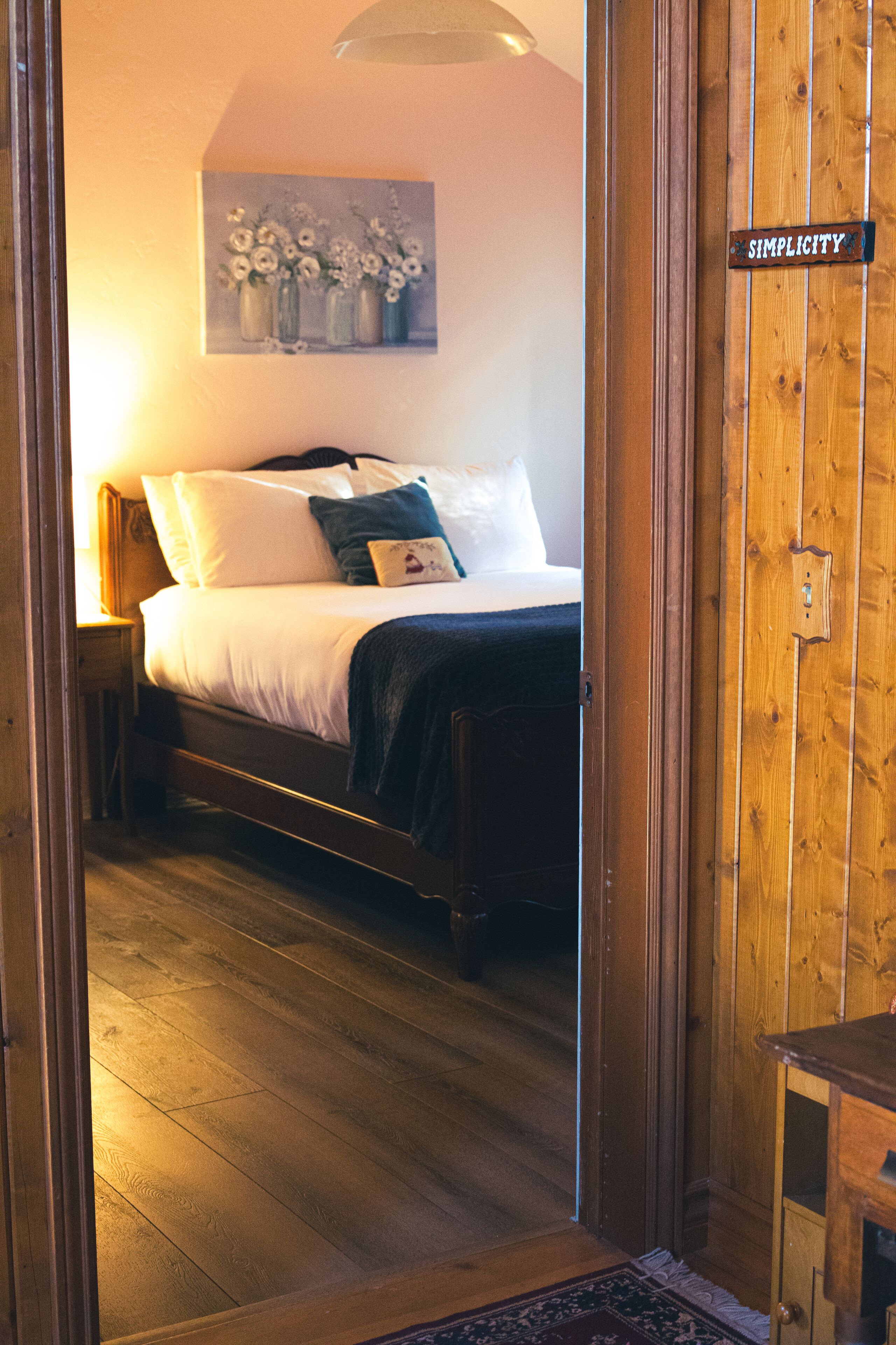 A bedroom with a bed, a nightstand, a painting on the wall, and a wooden door with a sign.