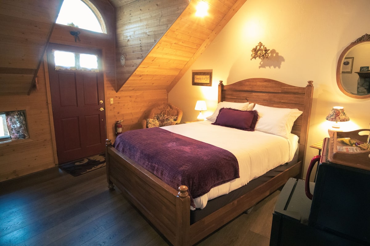 A wooden bedroom with a bed, a nightstand, a lamp, and a door.