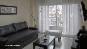 A living room with a black sofa, a small table, a white vase with branches, and a large window with white curtains. - Arenales Suites (Buenos Aires)
