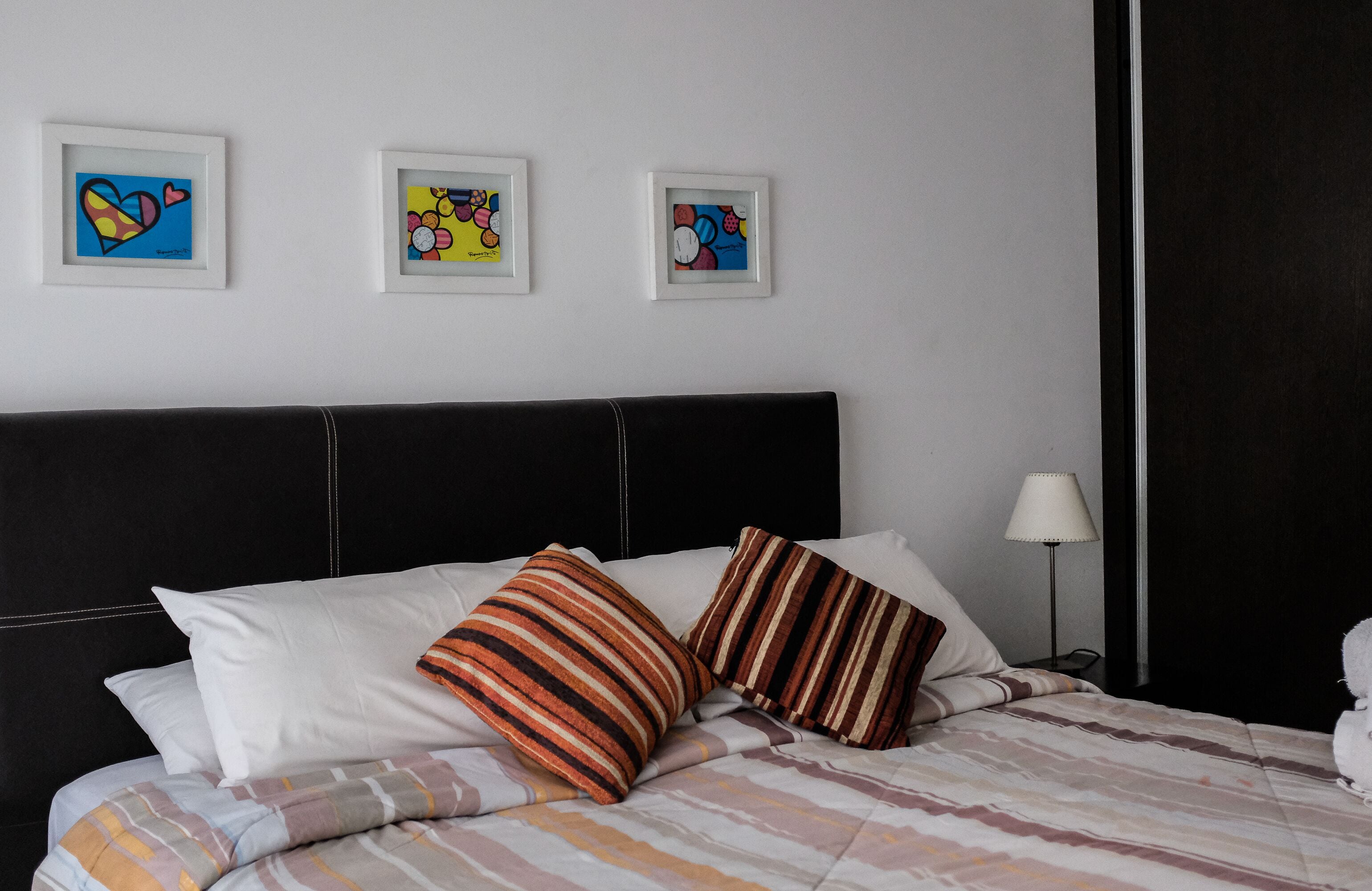 A neatly made bed with striped and solid pillows, a black headboard, and three colorful framed artworks on the wall.