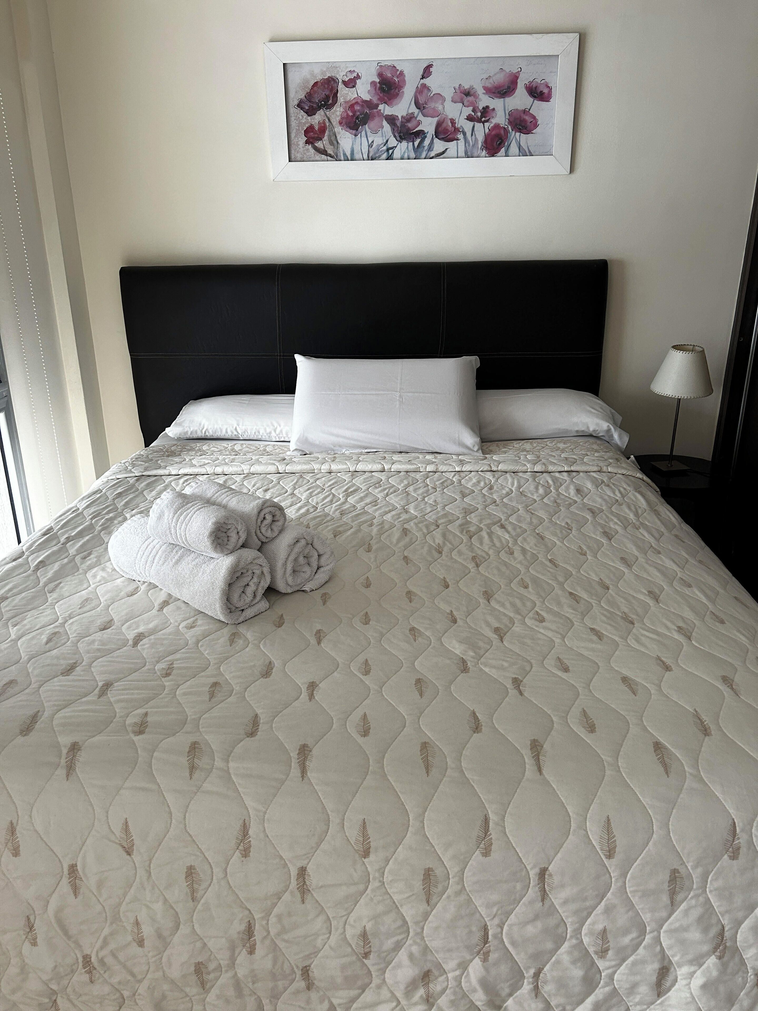 A neatly made bed with a quilted pattern and rolled towels, a black headboard, and a framed floral artwork on the wall.