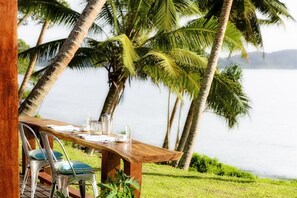 A wooden table set for two with metal chairs, positioned outdoors with palm trees and a body of water in the background.