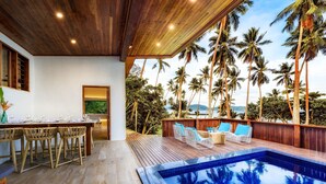 A poolside area with wooden flooring, a dining setup with chairs and a table, and a view of palm trees and a body of water. - The Remote Resort, Fiji Islands (Nawi)