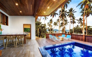 A poolside area with wooden flooring, a dining setup with chairs and a table, and a view of palm trees and a body of water.