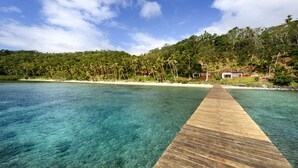 A wooden pier extending into a clear blue sea with a beach and lush greenery in the background. - The Remote Resort, Fiji Islands (Nawi)