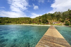 A wooden pier extending into a clear blue sea with a beach and lush greenery in the background.