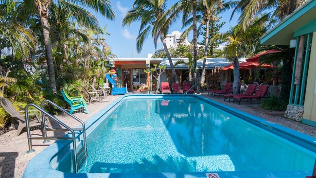 A swimming pool with lounge chairs and umbrellas, surrounded by palm trees and a building with a red awning. - Best Florida Resort (Lauderdale-by-the-Sea)