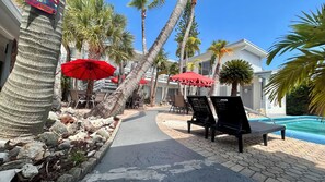 A poolside area with lounge chairs, red umbrellas, and palm trees.