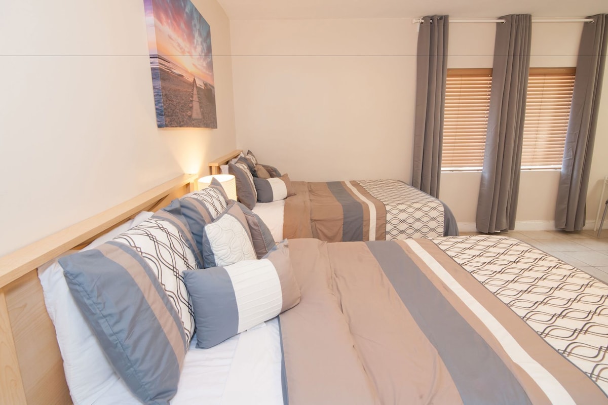 A bedroom with two beds, a headboard, a wall-mounted lamp, and a window with curtains.